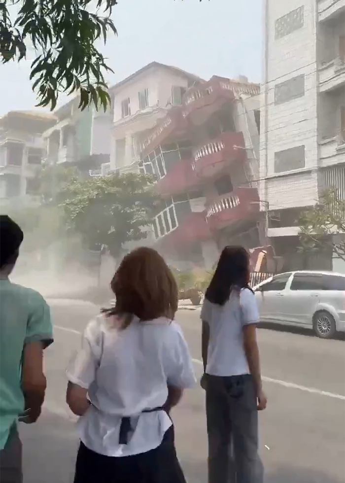 People witnessing the aftermath of a massive earthquake in Myanmar, with a collapsing building in the background.