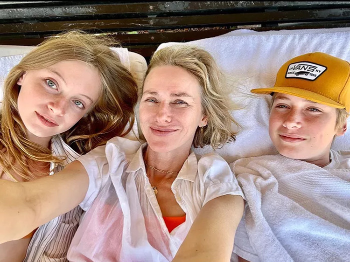 Naomi Watts with her two children, smiling while lying down together outdoors.