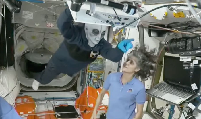 SpaceX crew member in costume on ISS interacting with NASA astronaut amidst Halloween-themed fun.
