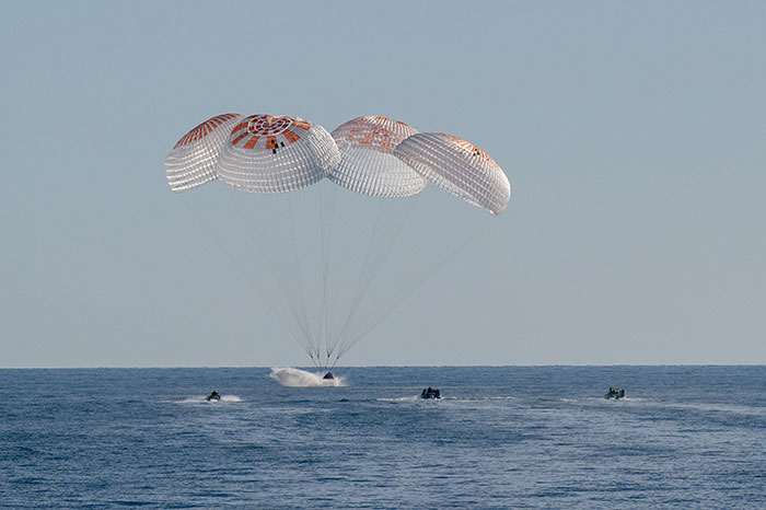 Parachutes open as astronauts splash down, with boats approaching for rescue.