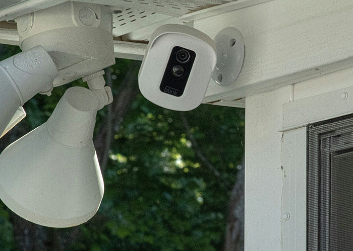 Security camera mounted under the roof, pointing towards house windows, with trees visible in the background. Security camera mounted under the roof, pointing towards house windows, with trees visible in the background.