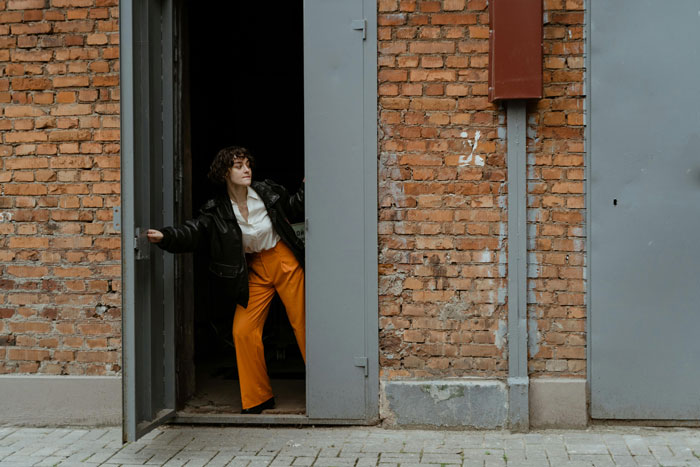 Woman in orange pants stepping out of a brick building through a gray door.