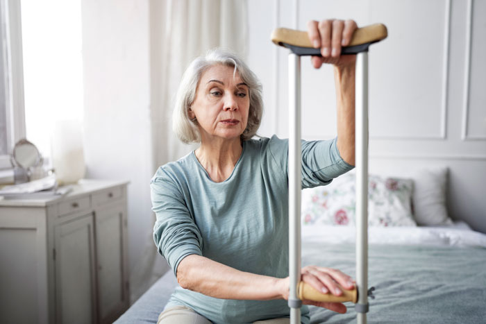 Elderly woman with crutch sitting on bed, reflecting on accessibility challenges.
