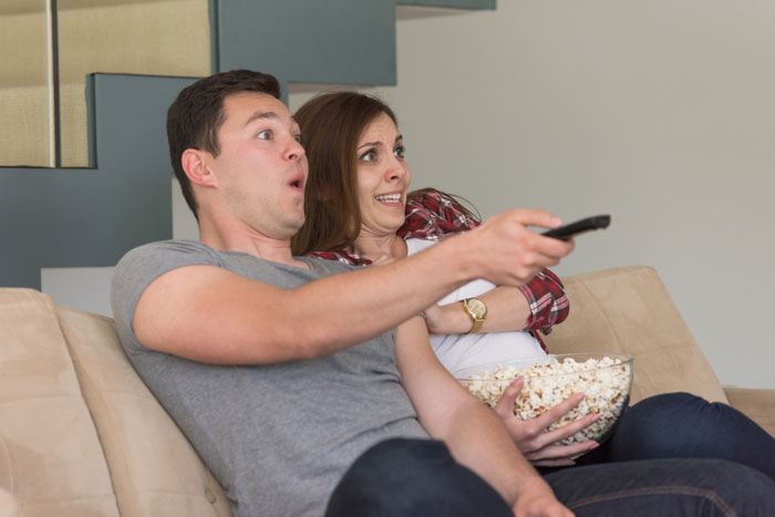 A surprised couple on the couch with popcorn, reacting to noise from neighbors treating the building like a nightclub.
