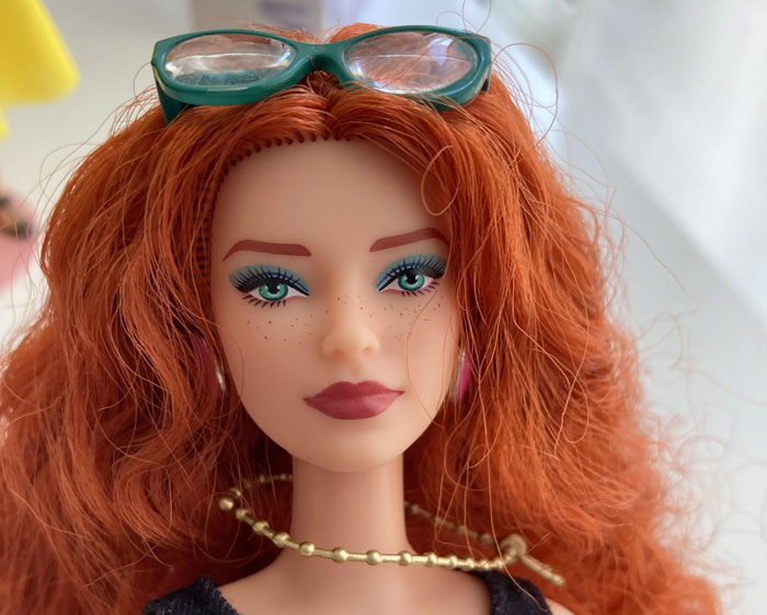 Customized doll with red hair, wearing green glasses and a gold necklace, symbolizing creativity and personalization.