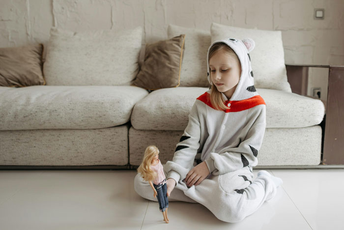 Child in animal onesie sitting on floor, holding a doll, by a couch.