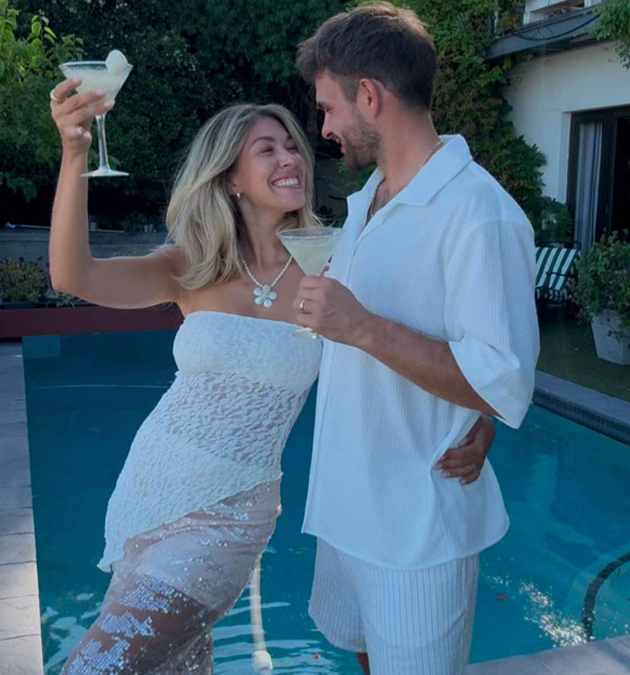 Bride smiling in a lace dress, holding a cocktail with groom by pool, showcasing a fun wedding moment.
