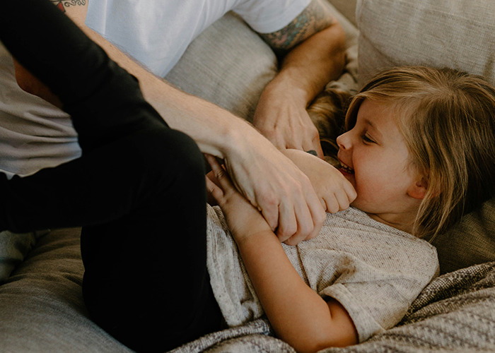 Child playfully tickling on a couch, smiling widely, with a joyful expression, capturing a moment from childhood experiences.