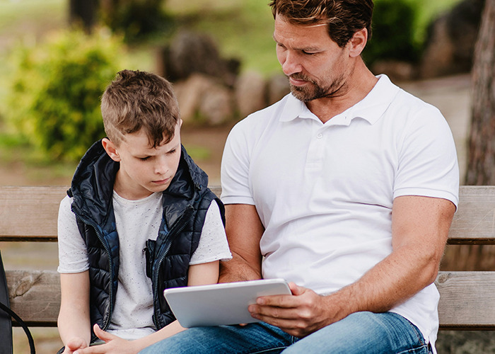 Father and son on a park bench, sharing a tablet and discussing childhood experiences.