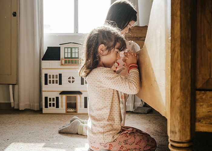 Children in pajamas kneeling and praying by a bed, with a dollhouse in the background, conveying childhood experiences.