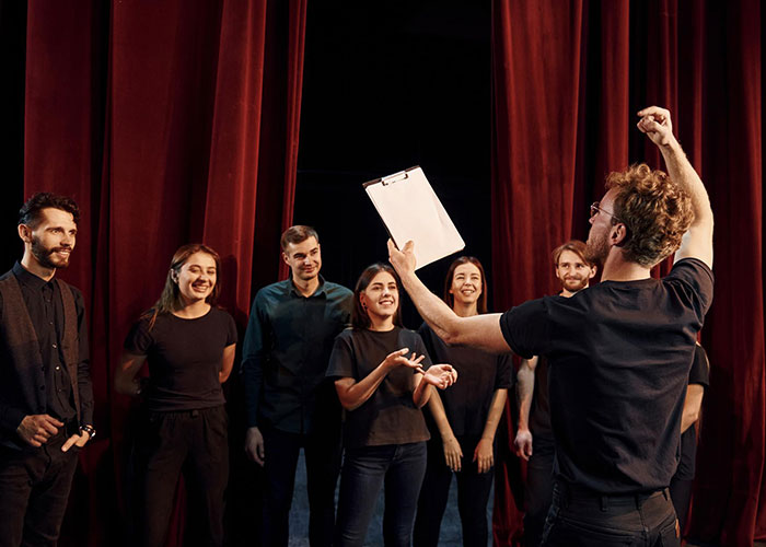 People in black clothing gather on stage, led by a man with a clipboard, resembling a cult-like gathering.