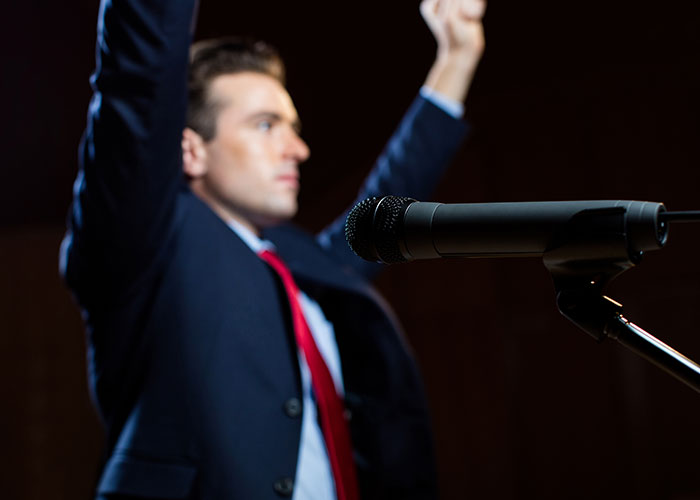 Man in a suit with raised arms near a microphone, suggesting a speech or presentation atmosphere.