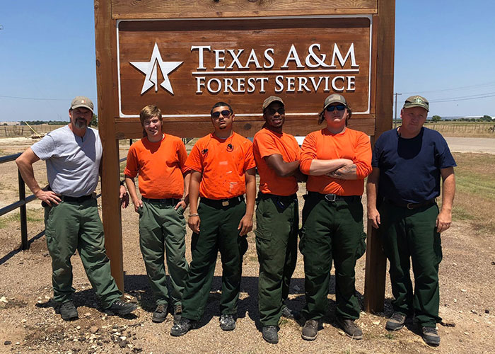 Group of six forestry workers in front of Texas A&M Forest Service sign, wearing orange and green uniforms.