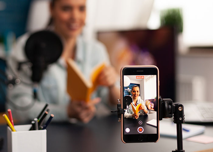 Person recording a video with a smartphone, holding a book, resembling mom groups' virtual activities.