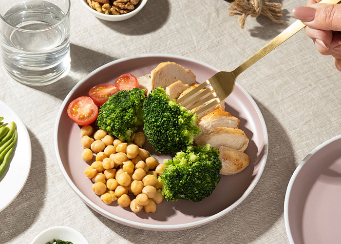 Plate of healthy food with broccoli, chicken, chickpeas, and tomatoes; person holding a fork over the meal.