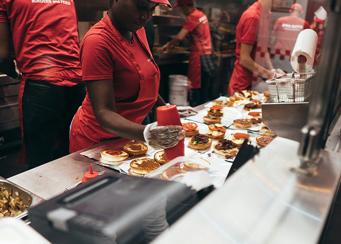 Worker in a red uniform preparing burgers in a busy kitchen, adding condiments to buns.