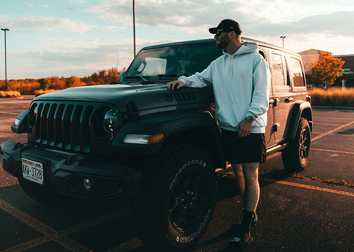 Man in a white hoodie and cap leaning on a Jeep in a parking lot during sunset.