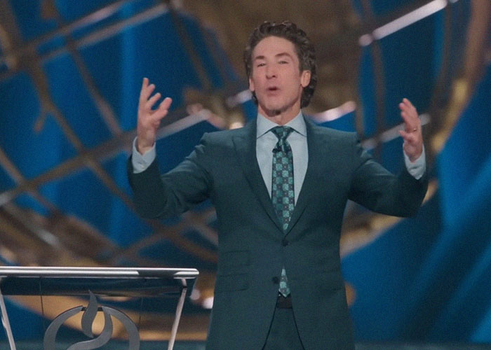 A person in a suit gestures passionately on a stage, lit by blue and gold lights, reminiscent of mom groups gatherings.