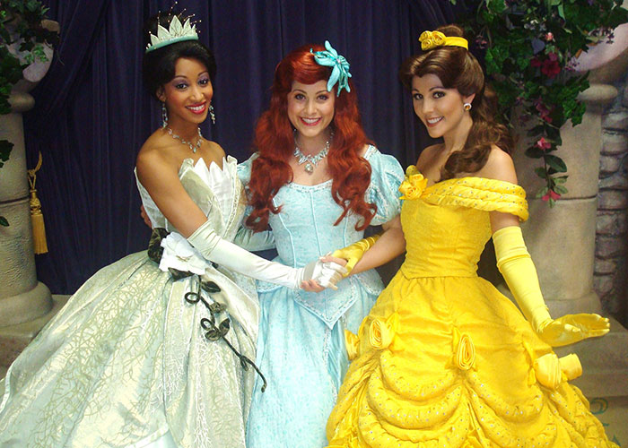 Three women dressed as princesses smiling and holding hands, evoking a playful mom groups vibe.