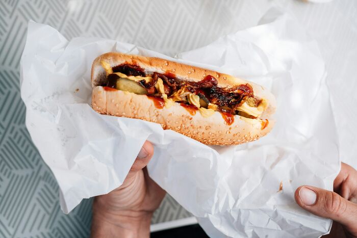 A person holds a fast food hot dog wrapped in white paper, topped with pickles and sauce.