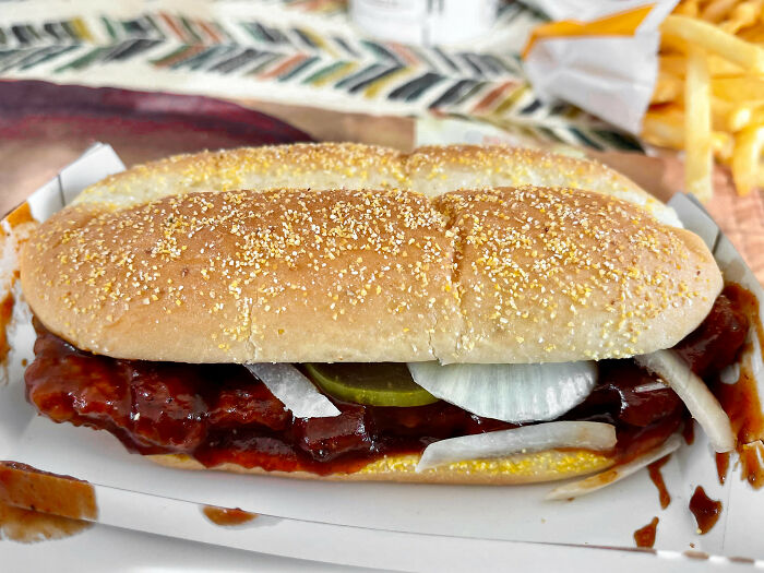 Hyped fast food sandwich with onions and pickles on a tray, next to a serving of fries in the background.