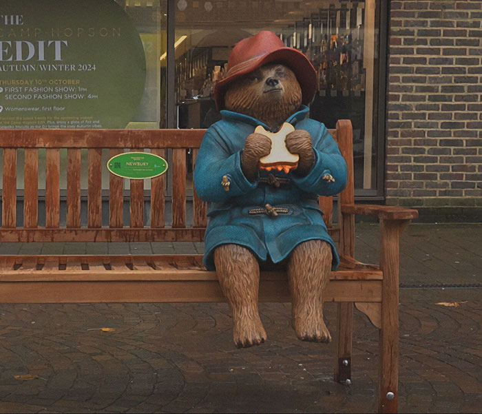Paddington Bear statue sitting on a bench, wearing a blue coat and red hat, in front of a shop display.