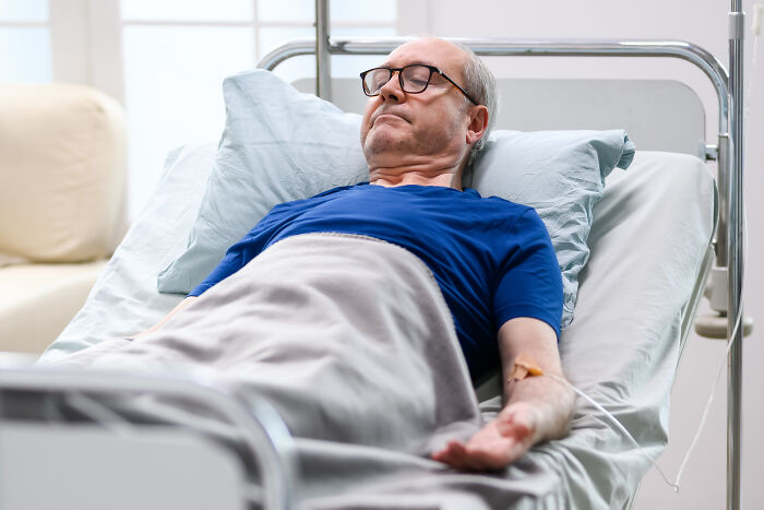 Elderly man in a hospital bed with an IV, wearing glasses and a blue shirt, representing an insignificant thing stuck forever.