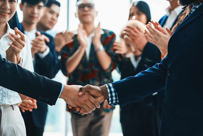 Older person and younger person shaking hands, surrounded by applauding people.