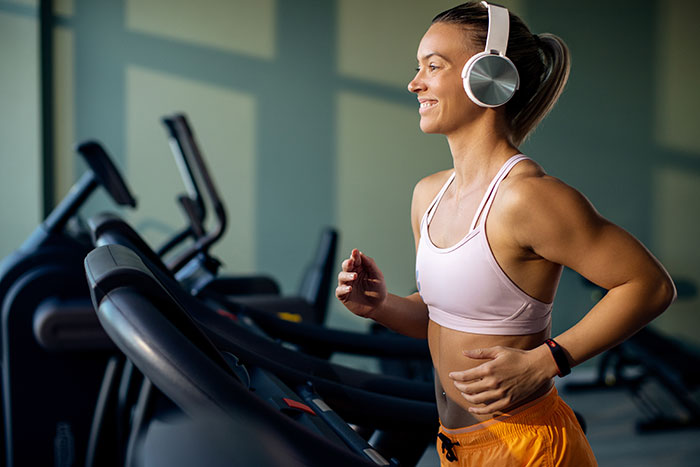 Woman jogging on treadmill in gym, wearing headphones, smiling confidently.