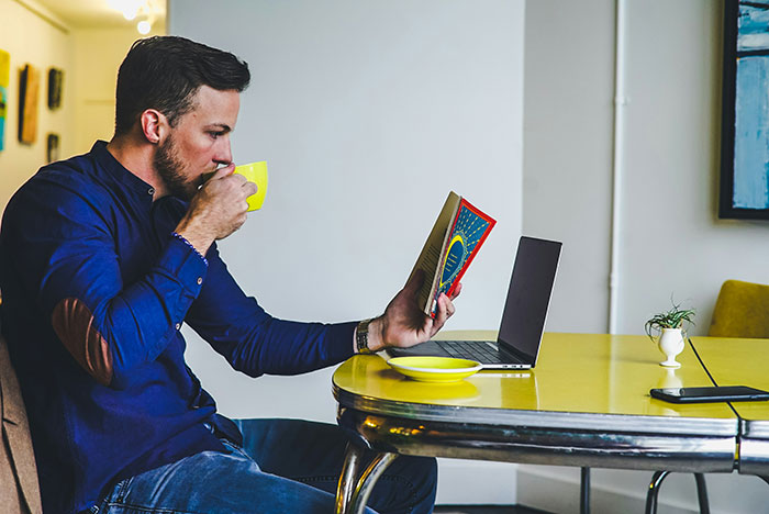 Man sipping coffee, reading a book at a table with a laptop; life insights from older people in focus.