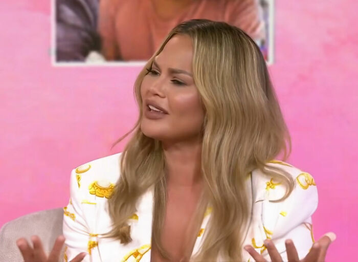 A woman with long blonde hair gestures while speaking, wearing a patterned white jacket in front of a pink background.