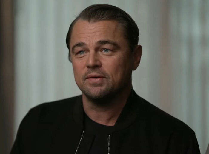 Man in a black jacket with slicked-back hair, expressing confidently during an interview; discusses perceptions of attractiveness.