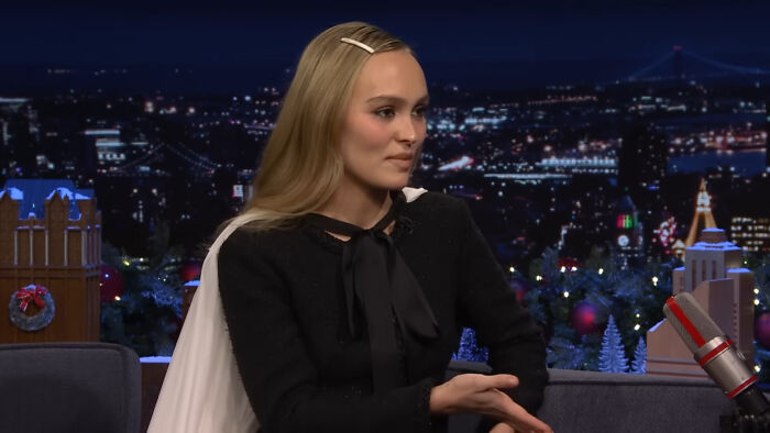 A person on a talk show set at night, discussing topics while gesturing with hands, in front of a scenic city backdrop.