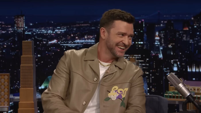 Man wearing a tan jacket on a talk show set, with a cityscape backdrop, discussing public perceptions of celebrity attractiveness.