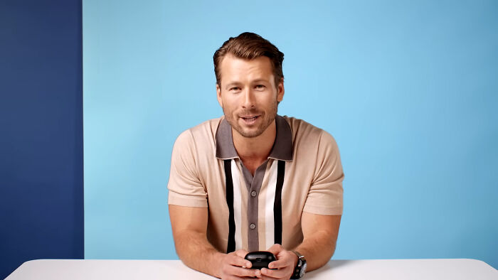 A man sitting at a table with a blue background, holding a device, related to celebrities and attractiveness opinions.