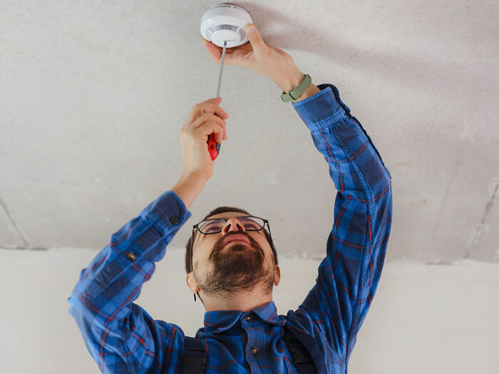 Man in blue plaid shirt fixing a smoke detector, illustrating home maintenance for first-time buyers.