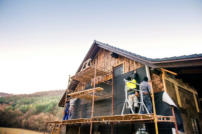 Construction workers on scaffolding renovating a first-time home amid scenic countryside.