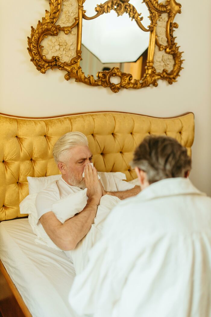 Elderly man sitting in a bed, raising hands to face, with another person beside him, under an ornate mirror.