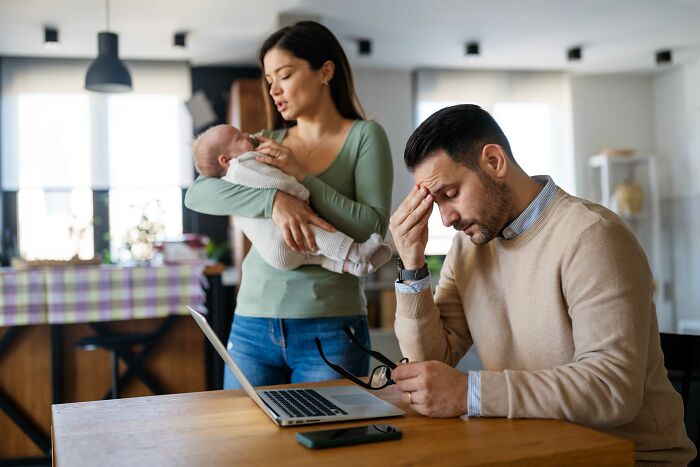 Man looking stressed at table with laptop, while a woman holds a baby nearby, illustrating decision not to have kids.