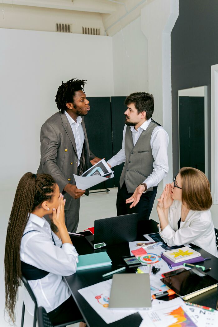 Office meeting with two men in suits discussing and two women seated at a table, embodying unprofessional work dynamics.
