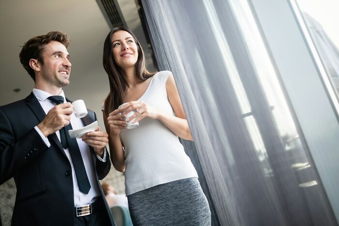 Two colleagues smiling, holding coffee cups, standing by a window, capturing a professional work moment.