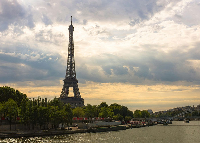 Eiffel Tower in Paris, France, with the Seine River in the foreground and a cloudy sky above.