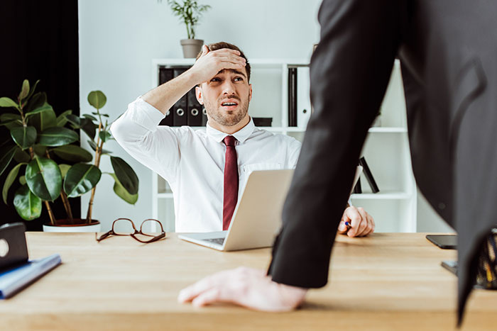 Employee looking stressed at a desk, next to a laptop, as a manager stands over them in an office setting.