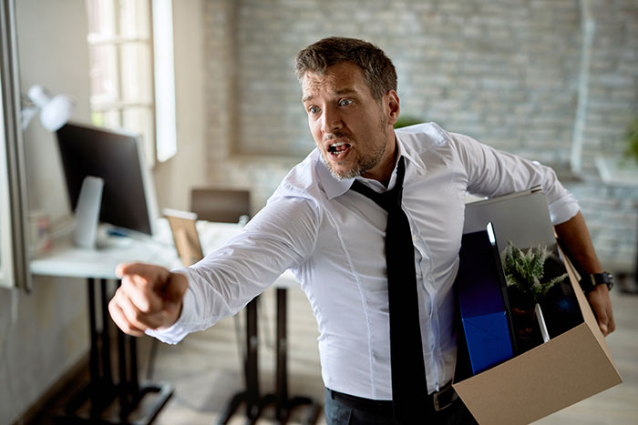 Man in office holding a box, pointing angrily; illustrates company firing scenario.