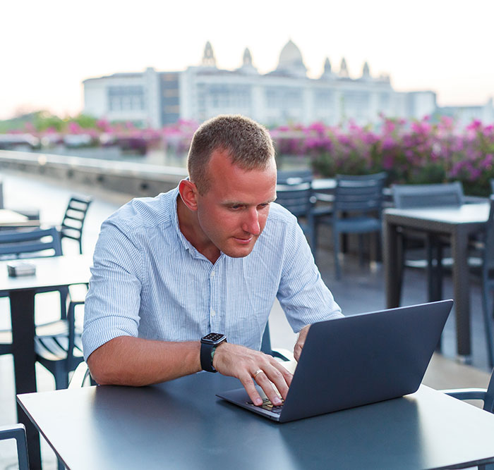 Man in blue shirt focusing on laptop while seated outdoors at a caf&eacute;, representing remote work challenges faced on vacation.