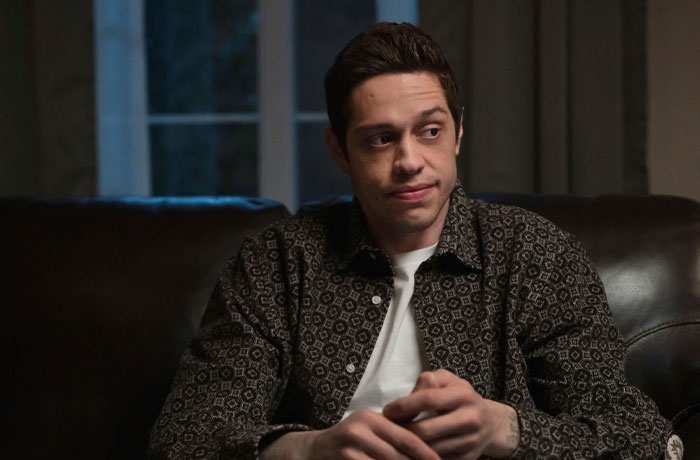 Pete Davidson sitting on a couch, wearing a patterned shirt, related to net worth and connections in entertainment.