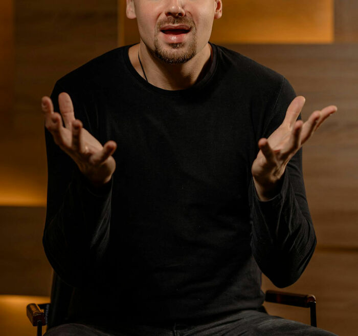 Man in a black shirt gesturing with open hands while seated, conveying strong emotions or disbelief.