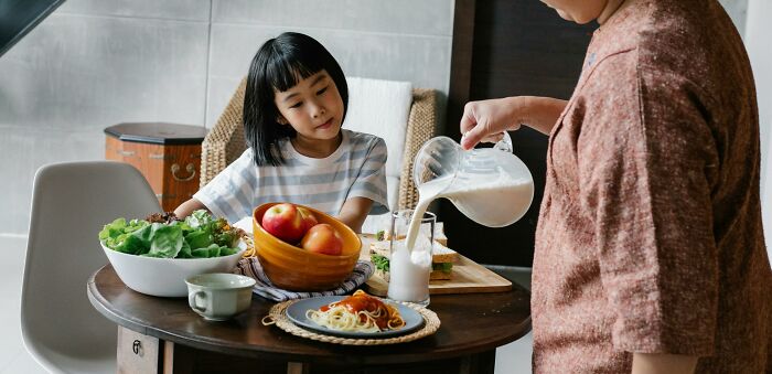 Child at table in middle-class home, observing as an adult pours milk, surrounded by a meal with salad and apples.
