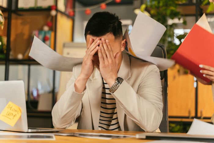 Frustrated office worker at desk, overwhelmed by overused expressions, holds head in hands amidst flying papers.