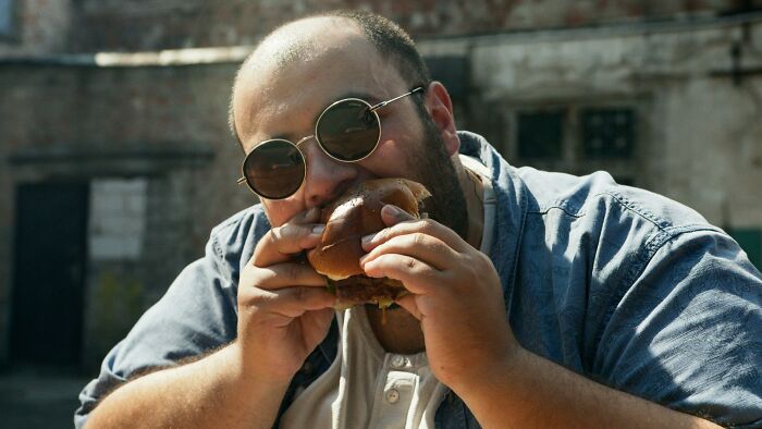 Man wearing sunglasses eating a burger outdoors, illustrating society's weird acceptance of certain habits.